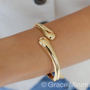 Könnte beinhalten: Ein goldfarbenes Armreif-Armband mit glatter, polierter Oberfläche. Das offene Armband hat abgerundete, längliche Enden. Der Schmuck wird an einem Arm vor dem Hintergrund eines weißen Hemdes und blauen Stoffes gezeigt.