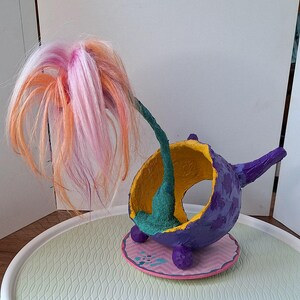 May include: A quirky, handmade sculpture. A purple bowl-shaped form with a yellow interior is supported by a teal stem and topped with pink, orange, and white faux hair. The sculpture sits on a pink and white patterned base with purple feet.