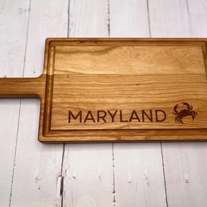 May include: A rectangular wooden cutting board with a handle. The board is light brown with a carved border and features the word "MARYLAND" and a crab silhouette. The handle has a hole for hanging.