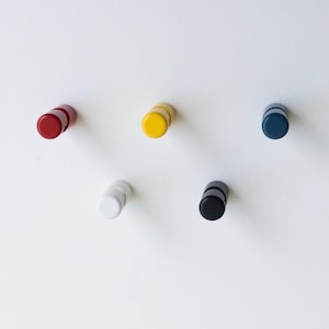Può includere: Cinque appendiabiti da parete colorati in rosso, giallo, bianco, nero e blu navy sono montati su un muro bianco. Ogni gancio ha una forma cilindrica con una faccia piatta e circolare. I ganci sono distanziati uniformemente.