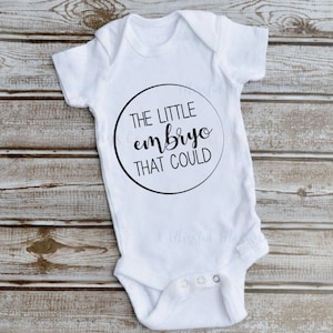Könnte beinhalten: Weißer Baby-Body mit dem Text "THE LITTLE embryo THAT COULD" in einem schwarzen Kreis. Der Body liegt auf einer Holzoberfläche. Die Worte "Blissful Blessings Co" stehen oben. Darunter steht "DIGITAL DOWNLOAD SVG - PNG".