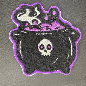 May include: A black patch featuring a cauldron design with a purple border. The cauldron has a skull on the front and is filled with purple liquid and white steam. This patch is suitable for Halloween or witch-themed crafts.