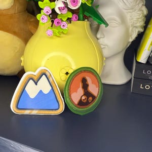 May include: Two decorative patches. One patch features a blue mountain design with a white peak and a gold border. The other patch is round with a silhouette and skull design, framed by a green border. Both patches are on a dark surface.
