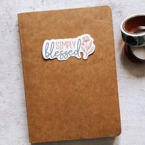 A brown notebook with a sticker that says "Simply Blessed" is placed on a white surface next to a roll of tape.