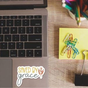 a laptop with a sticker that says "SAVED BY GRACE", a yellow sticky note with colorful pencils, and a black binder clip.