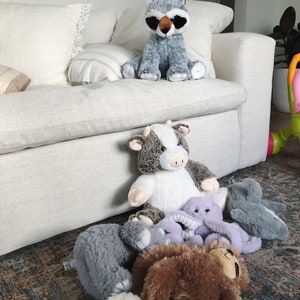 Puede incluir: Una colecci&oacute;n de peluches, incluyendo un mapache, una vaca, un pulpo, un perezoso y dos animales de peluche grises, est&aacute;n dispuestos en una alfombra frente a un sof&aacute; blanco.
