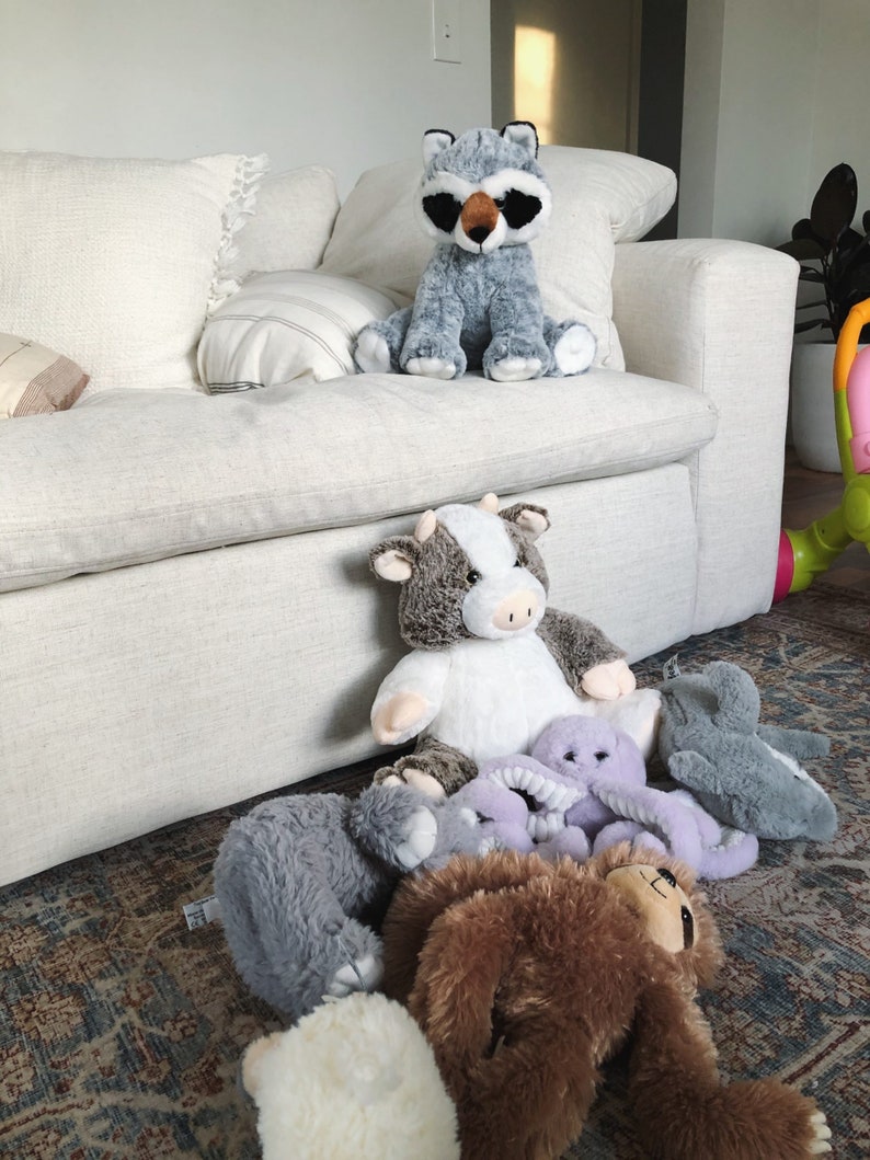 Puede incluir: Una colecci&oacute;n de peluches, incluyendo un mapache, una vaca, un pulpo, un perezoso y dos animales de peluche grises, est&aacute;n dispuestos en una alfombra frente a un sof&aacute; blanco.