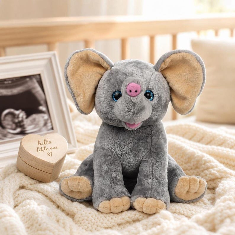 A gray stuffed elephant toy sits on a white blanket, with a wooden box and a framed photograph of a baby in the background.