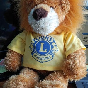 A brown teddy bear wearing a yellow t-shirt with a blue lion logo, sitting on a desk.