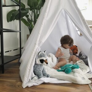 Puede incluir: Una tienda de campa&ntilde;a tipi blanca con un ni&ntilde;o sentado dentro jugando con animales de peluche. El ni&ntilde;o est&aacute; rodeado de una manta blanca y juguetes de peluche, incluyendo un mapache, un zorro, una tortuga y un animal de peluche gris.