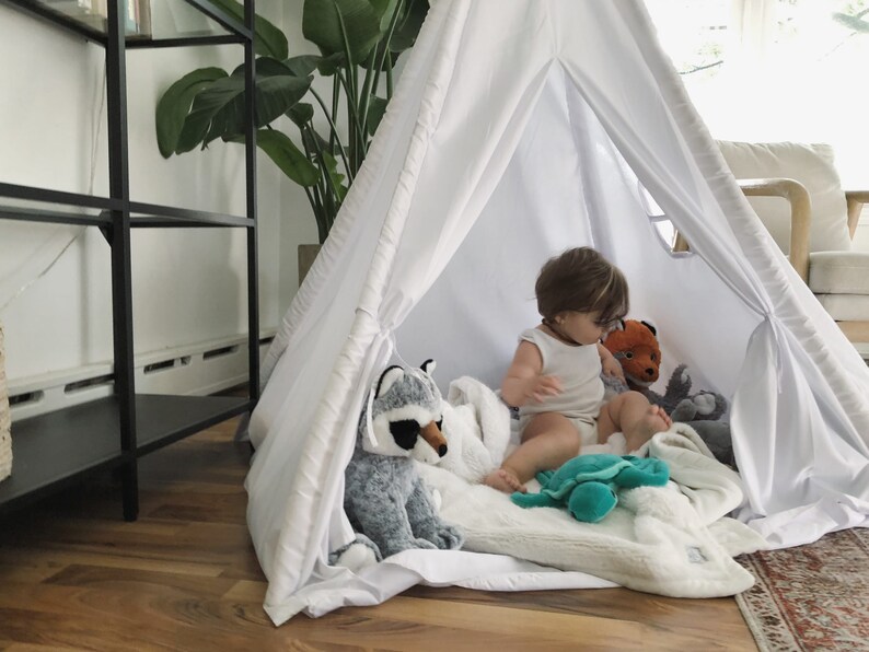 Puede incluir: Una tienda de campa&ntilde;a tipi blanca con un ni&ntilde;o sentado dentro jugando con animales de peluche. El ni&ntilde;o est&aacute; rodeado de una manta blanca y juguetes de peluche, incluyendo un mapache, un zorro, una tortuga y un animal de peluche gris.