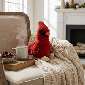 May include: A plush cardinal bird toy with bright red fur, a black face mask, and an orange beak. The stuffed animal is positioned next to a steaming ceramic mug, a small bowl of pinecones, and a thick book on a beige upholstered chair.