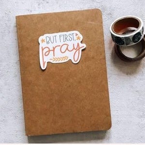 A brown notebook with a sticker that says "BUT FIRST PRAY" is placed on a white surface next to a roll of tape.