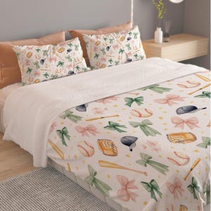 May include: A bedding set featuring a white comforter and two matching pillowcases decorated with a repeating pattern of baseballs, wooden bats, brown leather gloves, dark blue caps, and large pink and sage green bows on a cream background.