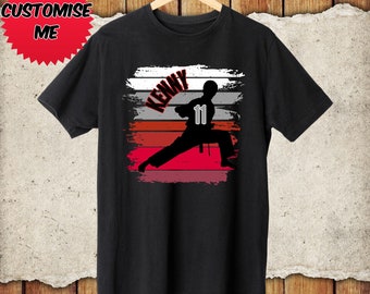 May include: Black t-shirt with a red, white, and gray striped background. A silhouette of a person in a karate pose is on the shirt. The name "KENNY" and the number "11" are on the shirt.