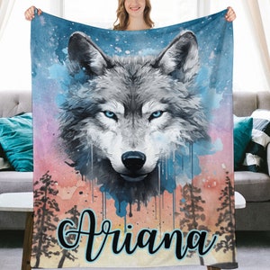May include: A soft, plush blanket featuring a watercolor painting of a wolf with blue eyes. The background is a colorful sunset with trees and the name "Ariana" is written in a blue script font.