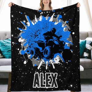May include: Black and blue personalized fleece blanket with a silhouette of a person riding an ATV in a blue paint splatter design. The name "ALEX" is printed in white at the bottom of the blanket.