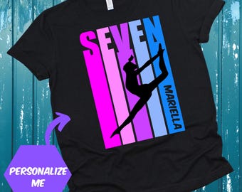 May include: Black t-shirt with a colorful retro design featuring the word "SEVEN" and a silhouette of a gymnast in a backbend pose. The name "MARIELLA" is printed vertically on the right side of the design.