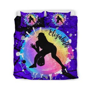 May include: A bedding set featuring a purple background with black splatter patterns. The center displays a colorful rainbow watercolor circle with a black silhouette of a softball player and the name Elizabeth in black script text.