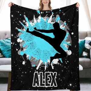 May include: Black and blue personalized fleece blanket with a silhouette of a person doing a martial arts kick in front of a blue and gray splatter paint design. The name "ALEX" is printed in white at the bottom of the blanket.
