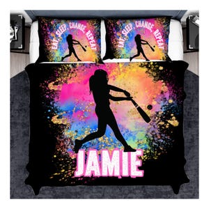May include: A black bedding set featuring a colorful paint splatter design with a silhouette of a softball player swinging a bat. The text "EAT SLEEP, CHANGE, REPEAT" appears on the pillowcases, and the name "JAMIE" is printed in large white letters on the comforter.