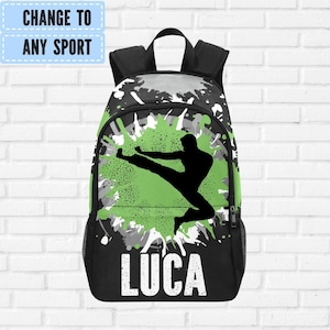 May include: Black backpack with a green and gray splatter design. A black silhouette of a person performing a martial arts kick is featured on the front. The name "LUCA" is printed in white block letters below the silhouette. The top of the backpack has a black handle.