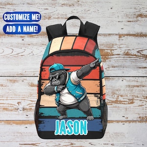 May include: A black backpack with a colorful retro design featuring a cartoon gorilla wearing sunglasses and a blue cap, dabbing. The name "JASON" is printed in blue. The text "CUSTOMIZE ME! ADD A NAME!" is also visible.