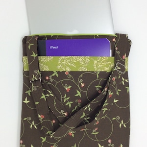 May include: A brown fabric tote bag with a green and white floral pattern. The bag has a zippered pocket on the front and a long strap. A laptop computer and a purple Mead notebook are inside the bag.