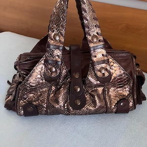 Op de afbeelding: Een donkerbruine handtas met een metallic bronskleurig slangenleer-textuurpaneel aan de voorzijde. De tas heeft twee gevlochten handvatten, donkerbruine lederen accenten en ronde metalen studs. Een centrale verticale lederen riem sluit de voorzijde.