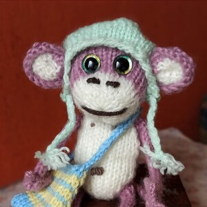 May include: A knitted purple monkey wearing a light blue and white hat and carrying a striped blue and yellow bag. The monkey is sitting on a wooden box.