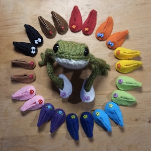 May include: A green knitted frog wearing white felt slippers with pink flowers. The frog is surrounded by a circle of felt slippers in various colours, including red, orange, yellow, green, blue, purple, and pink.