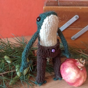 Puede incluir: Una rana verde tejida a mano con pantalones marrones y un botón morado. La rana está de pie sobre una superficie de madera con una rama de pino verde y una granada roja.