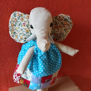 May include: A handmade stuffed elephant toy wearing a blue and white polka dot dress with a patchwork skirt. The elephant has floral fabric ears and black eyes.