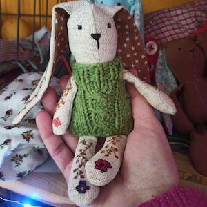 May include: A handmade plush bunny toy with brown and white ears, a green knitted sweater, and embroidered floral details on its arms and legs.