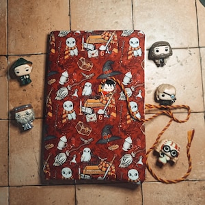 Puede incluir: Una libreta roja con un diseño de Harry Potter con búhos, pociones y el escudo de Hogwarts. La libreta está cerrada y tiene un cierre de cremallera. La libreta está rodeada de pequeñas figuras de personajes de Harry Potter.