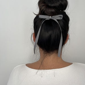May include: A grey ribbon bow hair accessory with a small pearl accent is tied in a bun. The ribbon hangs down the back of the head. The person is wearing a white long-sleeved top.