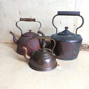 May include: Three metal tea kettles of varying sizes and finishes. One is a small, polished copper kettle, one is a medium-sized, weathered red kettle with a wooden handle, and one is a large, dark copper kettle with visible green oxidation.