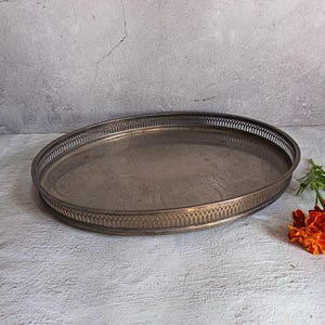 May include: An oval silver-toned metal serving tray featuring a decorative pierced gallery rim and an intricate engraved pattern on the flat base. The tray has a polished finish with visible signs of age and oxidation throughout the metal surface.