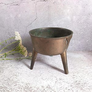 May include: A small, round, bronze-coloured metal bowl with a wide opening and three tapered legs. The interior surface shows signs of oxidation with visible green patina, while the exterior has a smooth, weathered metallic finish.