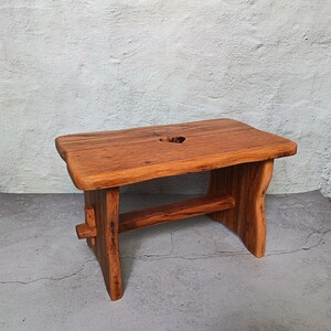 May include: A small, handcrafted wooden stool with a natural finish. The stool has a rectangular seat with a heart-shaped hole and sturdy legs connected by a horizontal support. The wood grain is visible, highlighting the natural beauty of the material.
