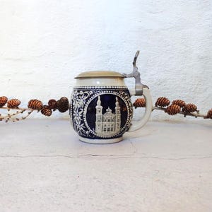 May include: A small, white ceramic beer stein with a blue and white design of a church. The stein has a metal lid and handle.
