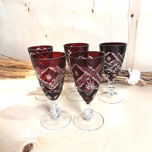 May include: A set of five stemmed glasses featuring deep red bowls with intricate cut-glass patterns, including star and diamond motifs. Each glass has a clear, transparent stem and a circular base, showcasing a contrast between the coloured glass.