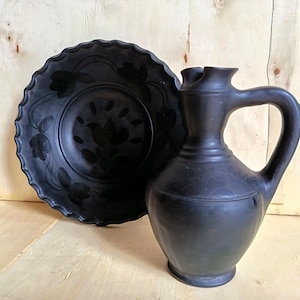 May include: A matte black ceramic pitcher with a curved handle and a wide spout, positioned next to a matching black ceramic plate featuring a scalloped edge and a subtle, dark floral pattern embossed on the surface of the round dish.