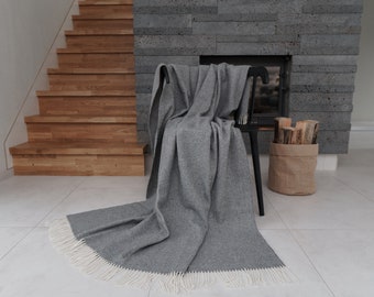 May include: A gray throw blanket with white fringe is draped over a black chair in front of a fireplace. The blanket is made of a soft, textured material and has a subtle herringbone pattern.