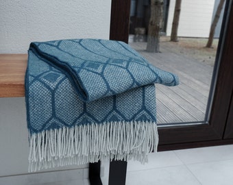 May include: A teal blue throw blanket with a geometric pattern and white fringe. The blanket is folded and placed on a wooden bench.