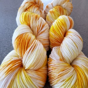 Puede incluir: Primer plano de varios ovillos de hilo teñido a mano. El hilo es una mezcla de blanco, amarillo y naranja, creando un efecto degradado. El hilo está bien enrollado y parece suave y esponjoso, ideal para proyectos de punto o ganchillo.