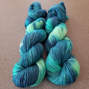 May include: Two skeins of hand-dyed yarn in shades of teal, turquoise, and seafoam green. The yarn is tightly wound, showing the color variations. Ideal for knitting or crochet projects.