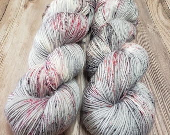 May include: Two skeins of speckled yarn in a light gray color with red and dark gray flecks. The yarn is arranged on a wooden surface, showcasing its texture and color variations. Ideal for knitting or crocheting projects.
