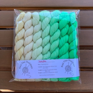 May include: A pack of Honeydew fingering/sock yarn in a gradient of colours from pale yellow to bright green. The package includes a label with the text "Knit The Holidays" and product details. The yarn is 80% Superwash Merino Wool and 20% Nylon.