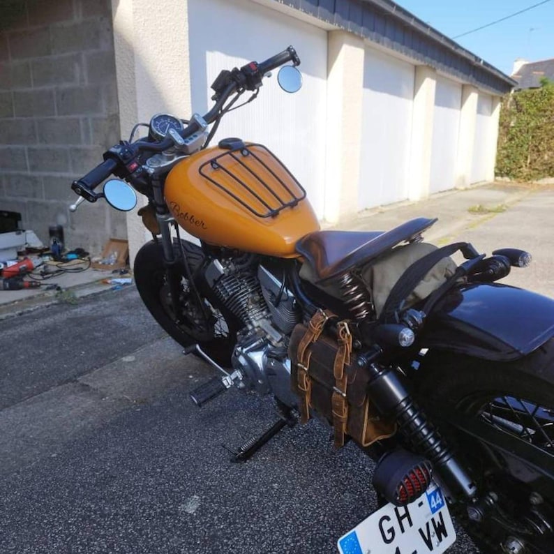 An orange motorcycle is parked outside on asphalt, fitted with a brown leather saddlebag on the side.